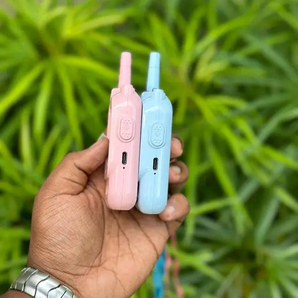 Hand holding two small electronic devices, one pink and one blue, against a green leafy background.